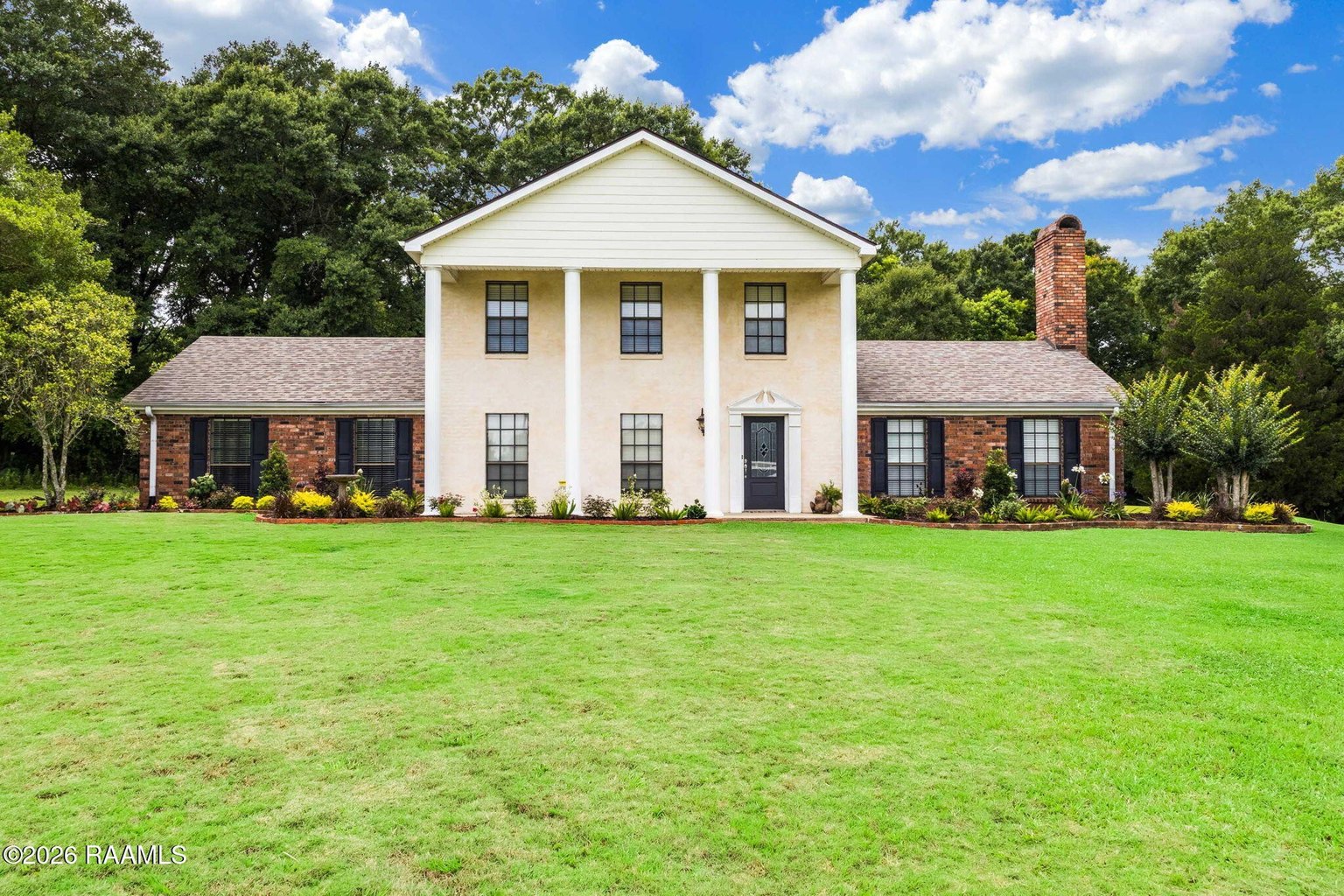 Front facade of two-story white and red brick colonial home with columned portico, gabled roof, brick chimney, landscaped beds, and manicured lawn.