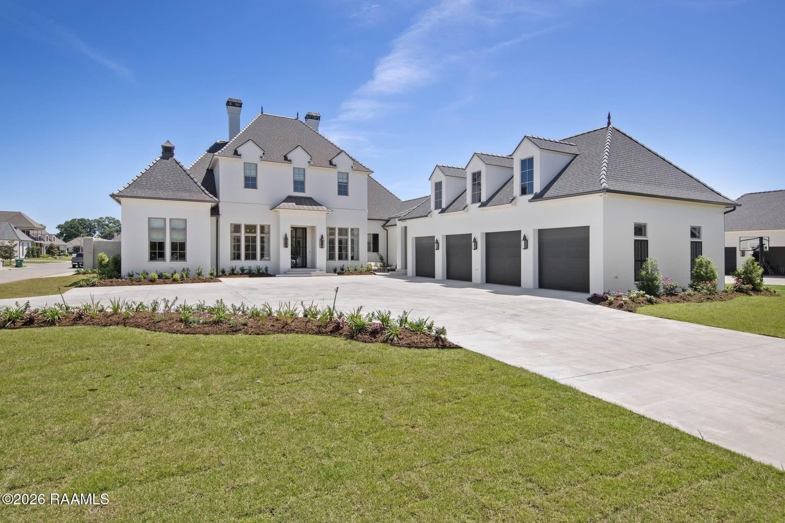 Angled front view of a two-story white brick French-style estate with a steep gray hipped roof, multiple dormers, copper cupolas, twin chimneys, an attached four-bay garage wing, and a wide concrete driveway framed by manicured lawn and landscaped flower beds under a clear blue sky.