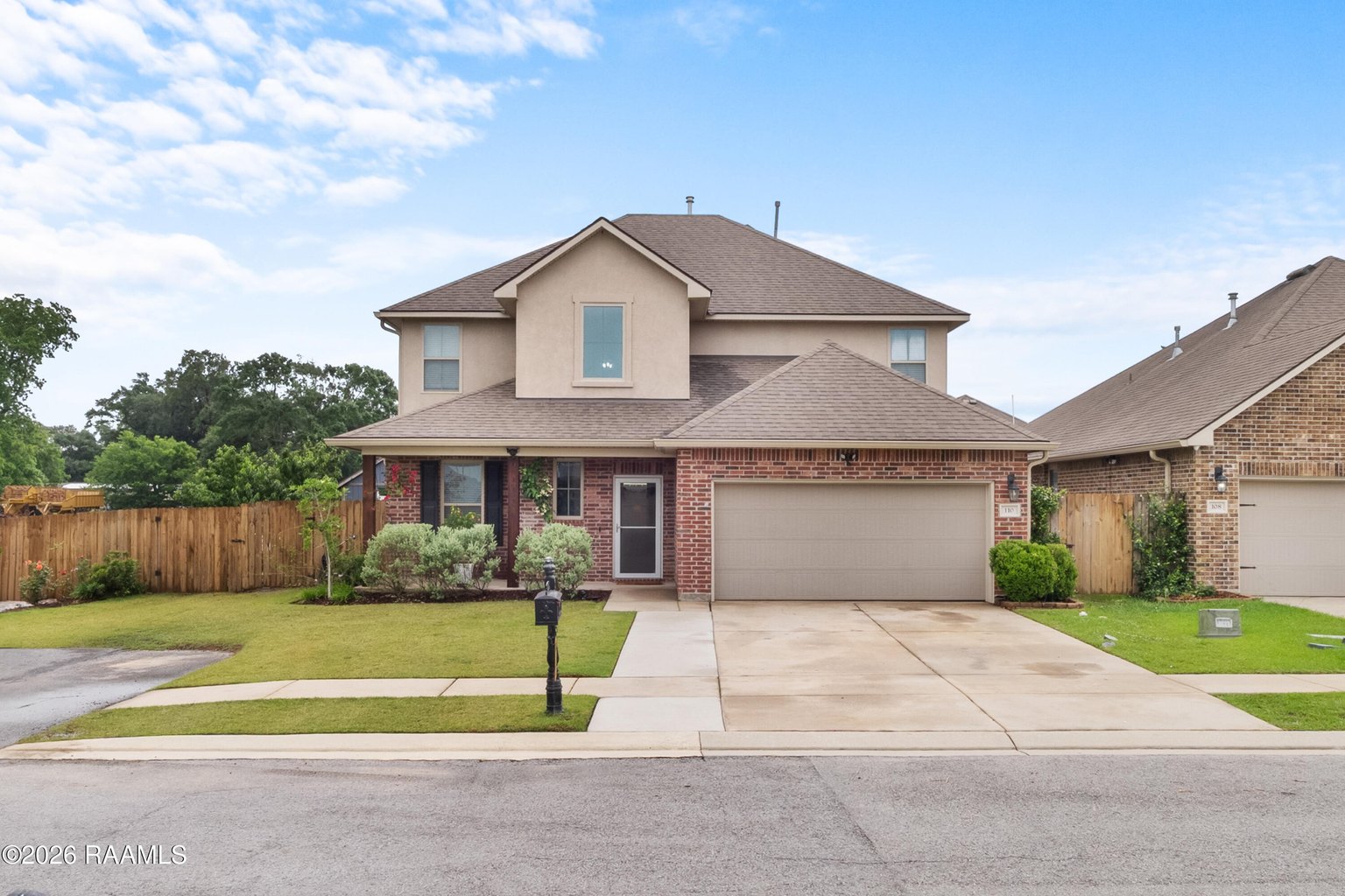 Brick two-story home with peaked roof, attached two-car garage, manicured lawn, wooden fence, black mailbox on grass strip, neighboring homes visible.