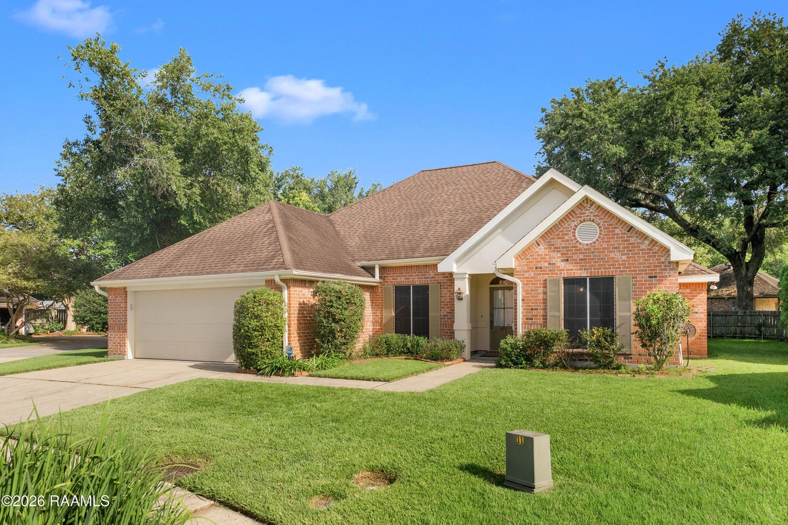 Red brick single-story home with white trim, two-car garage, arched entryway, landscaped front lawn with mature trees.
