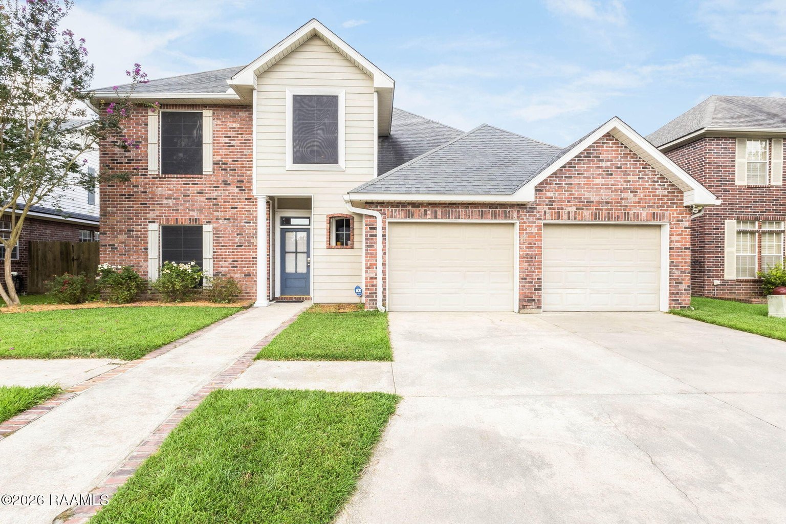 Two-story brick house exterior with cream vinyl siding, gabled roof, gray shingles, two two-car garage doors, front entry with dark door, landscaped front lawn bordered by brick walkway.