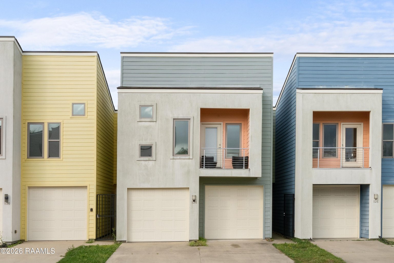 Exterior street view of modern connected townhouses with yellow, white, and blue siding, multiple stories, white garage doors, symmetrical window placement, concrete driveway.