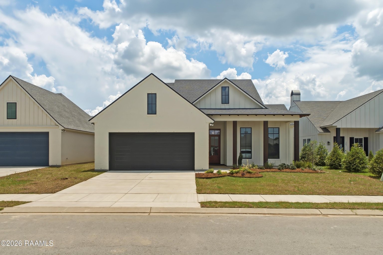 Front exterior of modern farmhouse with cream-colored siding, black two-car garage doors, gray pitched roof with dormers, white trim, concrete driveway, and landscape beds with shrubs.